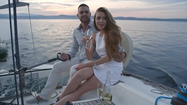 Handsome young man embracing his beautiful girlfriend while sailing on yacht at evening. They looking away and woman drinking wine