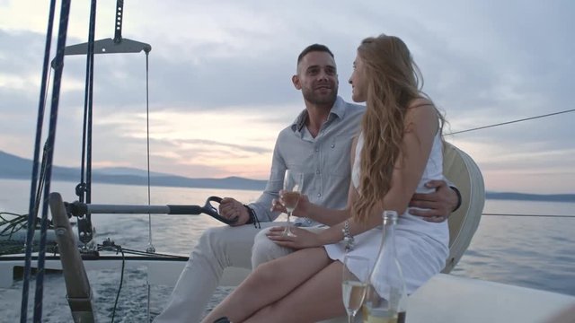 Handsome young man embracing his beautiful girlfriend while steering on yacht on lake. They talking and woman drinking wine