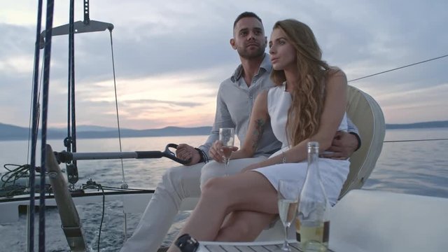 Romantic young couple sitting together on yacht and enjoying panoramic view of sunset. Girl holding wine glass and man steering yacht