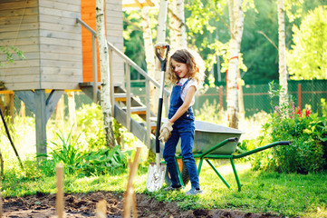 happy kid girl playing in summer garden with wheelbarrow and shovel and helping