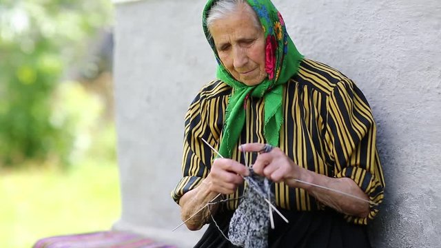 Ukrainian old woman is engaged in knitting. Old woman sits on bench and knits woollen socks. Senior woman in green headscarf sits near his house and knits sock. Female in kerchief knits woollen sock