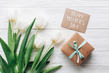 Obraz na płótnie Canvas happy mothers day text sign on stylish craft present box and greeting card and tulips on white wooden rustic background. flat lay with flowers empty paper with space for text. holiday gift
