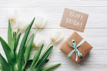 Obraz na płótnie Canvas happy birthday text sign on stylish craft present box and greeting card and tulips on white wooden rustic background. flat lay with flowers empty paper with space for text. holiday gift