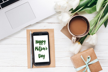 happy mothers day text sign on phone screen and laptop with morning coffee and tulips on white wooden rustic background. stylish flat lay with flowers and working gadgets with space for text.