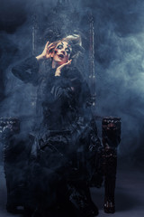 Young beautiful witch sits on a chair. Bright make up, skull, smoke-  halloween theme.