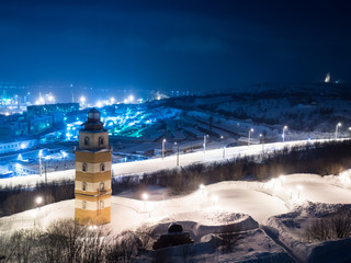 Night image of the city of Murmansk and the Memorial Complex of the sailors who died in peacetime with beacon tower against the backdrop of the sea port and snow covered streets and Alyosha monument