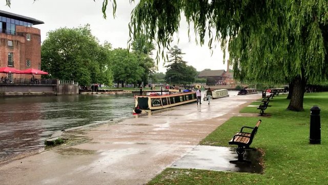 People walking alongside river in park on rainy day in summer  wet sidewalk with benches and willow trees - Stratford-upon-Avon  UK