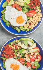 Close-up of healthy breakfast with fried egg, chickpea sprouts, seeds, vegetables and greens in bowls over grey concrete background, top view. Clean eating, healthy lifestyle, vegetarian food concept