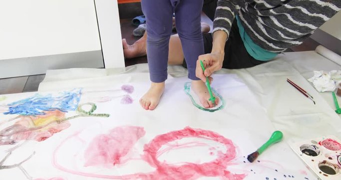 mother with brush painting outlining foot of three years barefoot child with blue pants, on white paper in the floor
