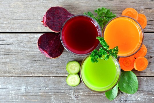 Three glasses of healthy vegetable juice with surrounding ingredients, above view over rustic wood