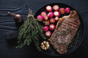Barbecued striploin steak and roasted radish in a frying pan over black wooden background, top view