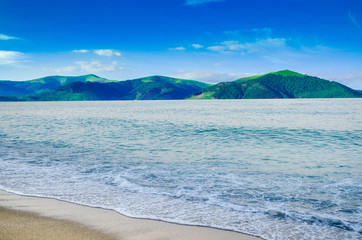summer landscape, the sand on the beach, blue sea with waves. Green mountains in the background of the sea