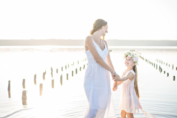 Fototapeta na wymiar Mom and daughter in white dresses with garlands of flowers on her head at sunset