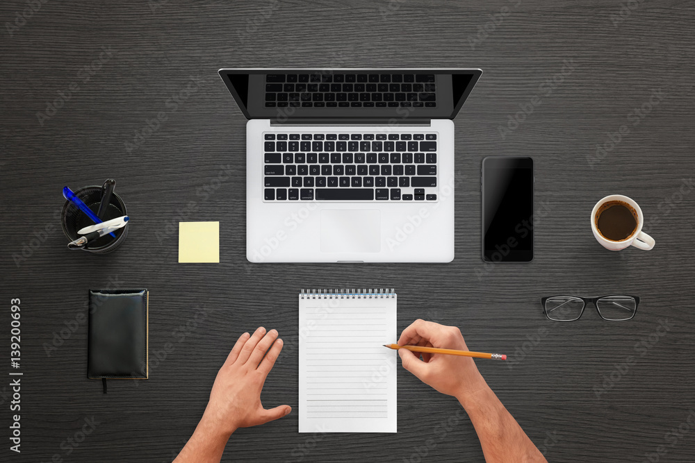 Poster Man write on pad. Top view of work desk with laptop, mobile phone with blank display screen for mockup. Glasses, cup of coffee, pens beside. Black wooden table texture in background.