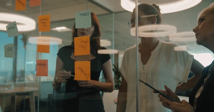 Three business women having a meeting in office. They are standing in front of glass wall with post it notes.