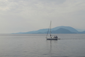 Fototapeta na wymiar Grey misty day, tranquil sea with smooth surface, yacht in the calm water, silhouette of an island on the horizon