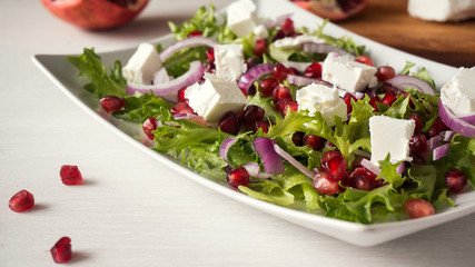 Fresh spring salad with feta cheese, red onion and pomegranate seeds in white dish on wooden background