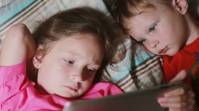 Girl and boy lying on the bed and watching cartoon on touchscreen tablet. Brother and sister having rest together.