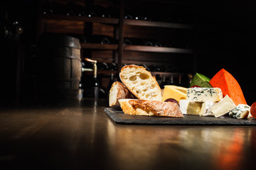 Mixed cheese lies on black plate before shelves with bottles of wine