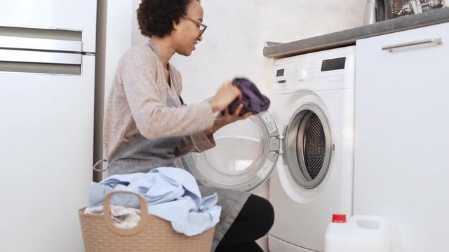 Young dark skinned female in apron and glasses loading clothes in washing machine from flasketin slowmotion