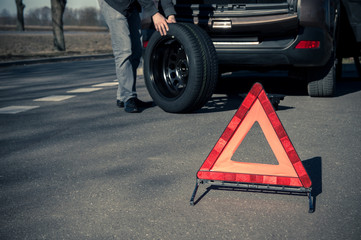 Warning triangle stands on the tarmac road and man holds the spare tire on the background