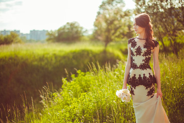 Shiny young woman in beige dress with black laces stands in tall grass