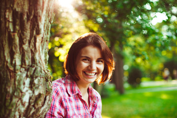 Fototapeta premium Charming girl in pink plaid shirt leans to old tree in park