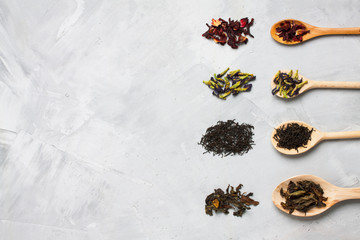 Wooden spoons with different tea leaves on grey concrete background.