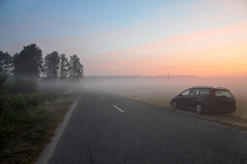 Asphalt road with a parked car in the morning mist on the background of dawn.