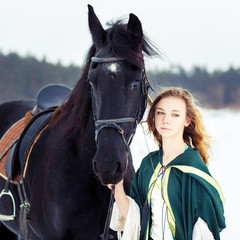 Young girl in white dress and green cape with black thoroughbred horse on winter field. Historical image