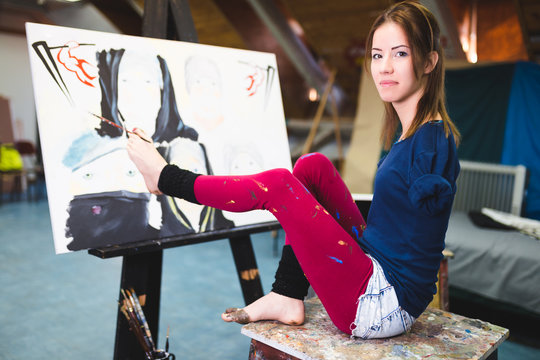 Disabled beautiful young artist painting incredible scenes in attic by holding paintbrush in her toes.