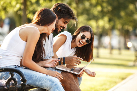 Young people at the park spending time together.They sitting on a wooden bench and using a laptop, friendship concept.