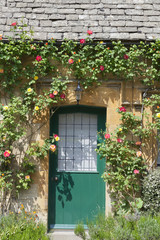 Obraz premium Green wooden and glass front doors in an old traditional English lime stone cottage surrounded by climbing red roses, lavender, on summer day .