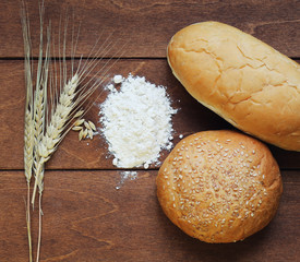 bun and a loaf of white bread, flour and wheat ears lying on old wooden table