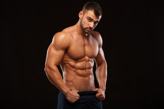 Strong Athletic Man - Fitness Model showing Torso with six pack abs. stands straight and puts his hands in trousers. isolated on black background with copyspace.