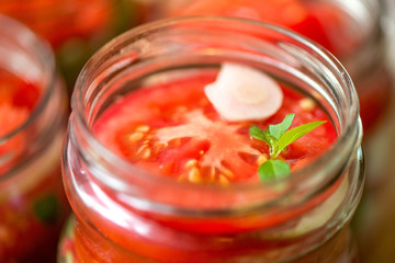 Fototapeta na wymiar Canning fresh tomatoes with onions for winter in jelly marinade. Macro shot of basil leaves on top of a red ripe tomato slice being put in jar.