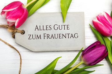 Women's day card with German words 'Alles gute zum frauentag' - All the best for women's day.Tulip flower and small heart on white wooden background.