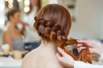Beautiful red-haired hairy girl, hairdresser weaves a French braid, close-up in a beauty salon....