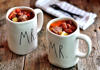 Delicious soup of beef meat with onions, potatoes, carrots, and red beans in cups on the old wooden background.