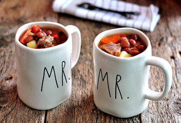 Delicious soup of beef meat with onions, potatoes, carrots, and red beans in cups on the old wooden background.