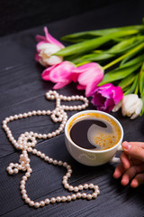 Bouquet of tender pink tulips and hands holding cup of coffee and pearls on black wooden background