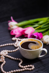Cup of coffee, pearl necklace and bouquet of tulips on black wooden background