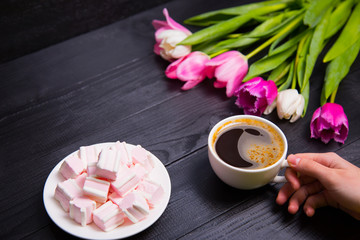 Bouquet of tender pink tulips and hands holding cup of coffee and marshmallows on black wooden background