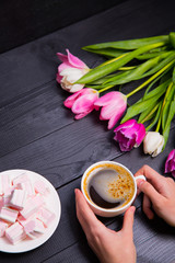 Bouquet of tender pink tulips and hands holding cup of coffee and marshmallows on black wooden background