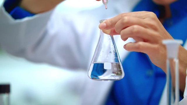 Woman chemist adding chemical reagent in pure liquid at glass flask. Close up of scientist hands put lab flask on table