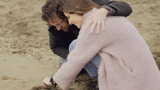 Beautiful woman painting heart on sand hugged by loving boyfriend closeup