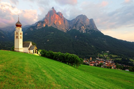 Sunset scenery of Church St. Valentin on green grassy hillside, rugged peaks of Mountain Schlern with alpenglow in background & Village Seis am Schlern in the valley in South Tyrol, Italy, Europe