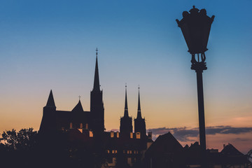 Wroclaw Cathedral island view from Oder river by sunset. Holy Cross church, St. John the Baptist cathedral and street lantern silhouettes by golden hour. Cathedral island in Wroclaw, Poland.