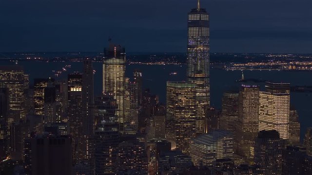 AERIAL HELI SHOT, CLOSE UP: Flying toward famous landmarked skyscrapers lit up with lights after the sunset in Lower Manhattan business district. Waterfront office buildings and New York Upper Bay