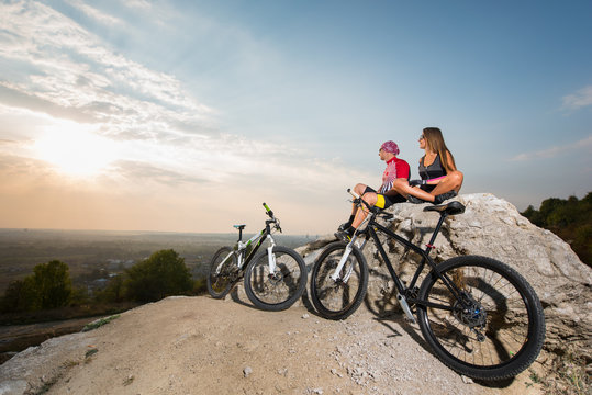 Cyclist couple in the sunglasses sitting on a rock near bikes, looking to the sun. On the background of forest, blue sky and sunset. Adventure travel in mountains on bike